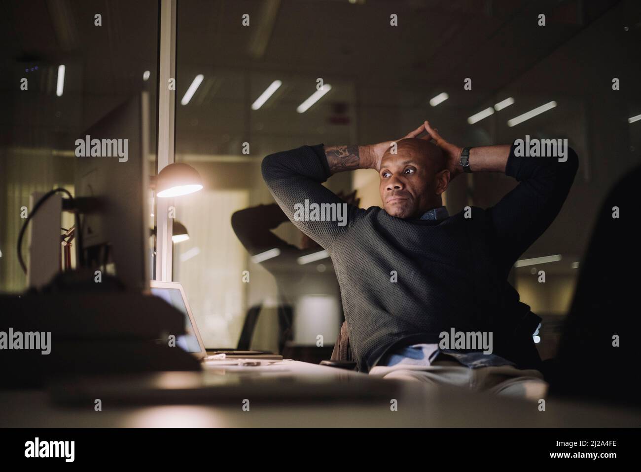 Ein reifer Geschäftsmann mit den Händen hinter dem Kopf, der spät in der Nacht im Büro arbeitet Stockfoto