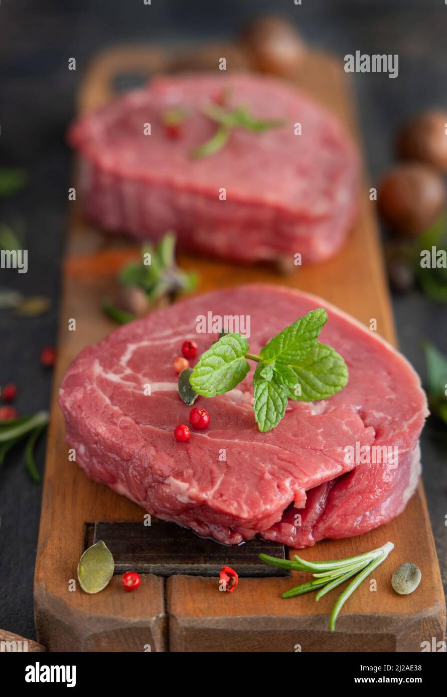 Rohe Rinderfiletsteaks auf Holzbrett auf dunklem Hintergrund Nahaufnahme. Portionierte Filet-Mignons mit Gewürzen und Kräutern. Konzept der Sommerkost Stockfoto