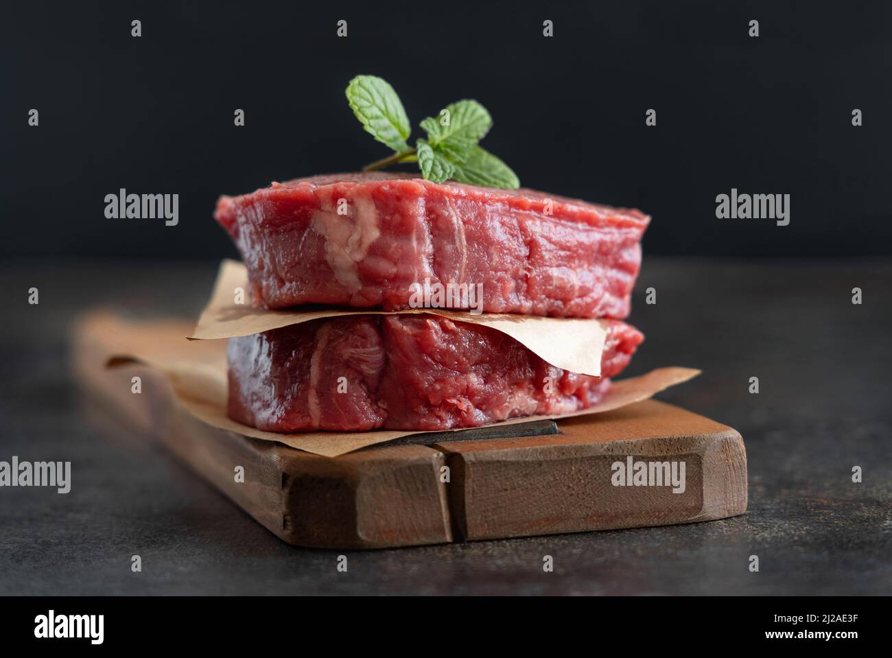 Rohe Rinderfiletsteaks auf Holzbrett auf dunklem Hintergrund Nahaufnahme. Portionierte Filet-Mignons mit Gewürzen und Zutaten. Konzept der Sommerkost Stockfoto