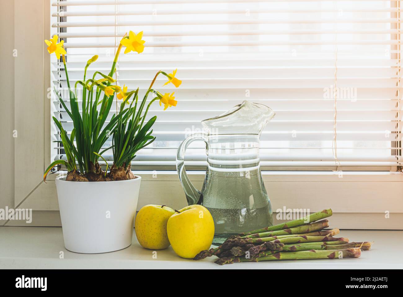 Frühlingsblumen Narzissen in Blumentopf, frische grüne Äpfel und Spargel auf Fensterbank. Konzept von Bioprodukten, gesunde Ernährung, Gartenarbeit Stockfoto