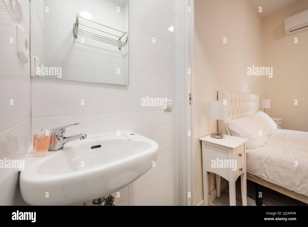 Badezimmer mit Wandwaschbecken aus Porzellan, quadratischem rahmenlosem Spiegel und Schlafzimmer mit weißer Bettwäsche und weißen Nachttischen Stockfoto