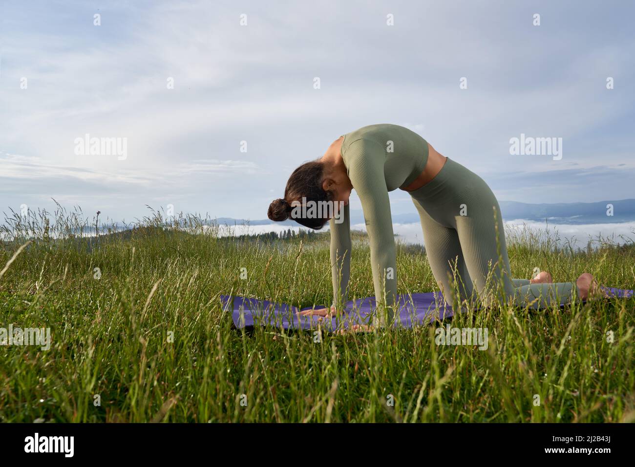 Charmante junge Frau in grüner Sportkleidung, die Yoga-Übungen an der frischen Luft macht. Aktive Brünette genießen Outdoor-Training unter den Sommerbergen. Stockfoto