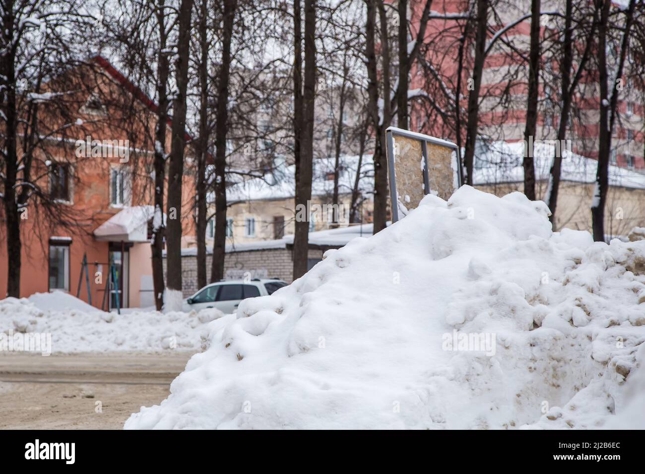 Schmutzige Schneeverwehung an der Straße vor dem Hintergrund von Stadthäusern und Bäumen. Auf der Straße liegt schmutziger Schnee in hohen Haufen. Urbane Winterlandschaft. Wolkiger Wintertag, weiches Licht. Stockfoto