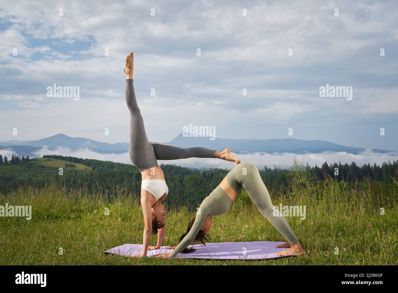 Zwei kaukasische Frauen in Sportkleidung üben während des Sommers unter den Bergen Akro-Yoga. Konzept der Menschen, gesunde Lebensstile und Outdoor-Aktivitäten. Stockfoto