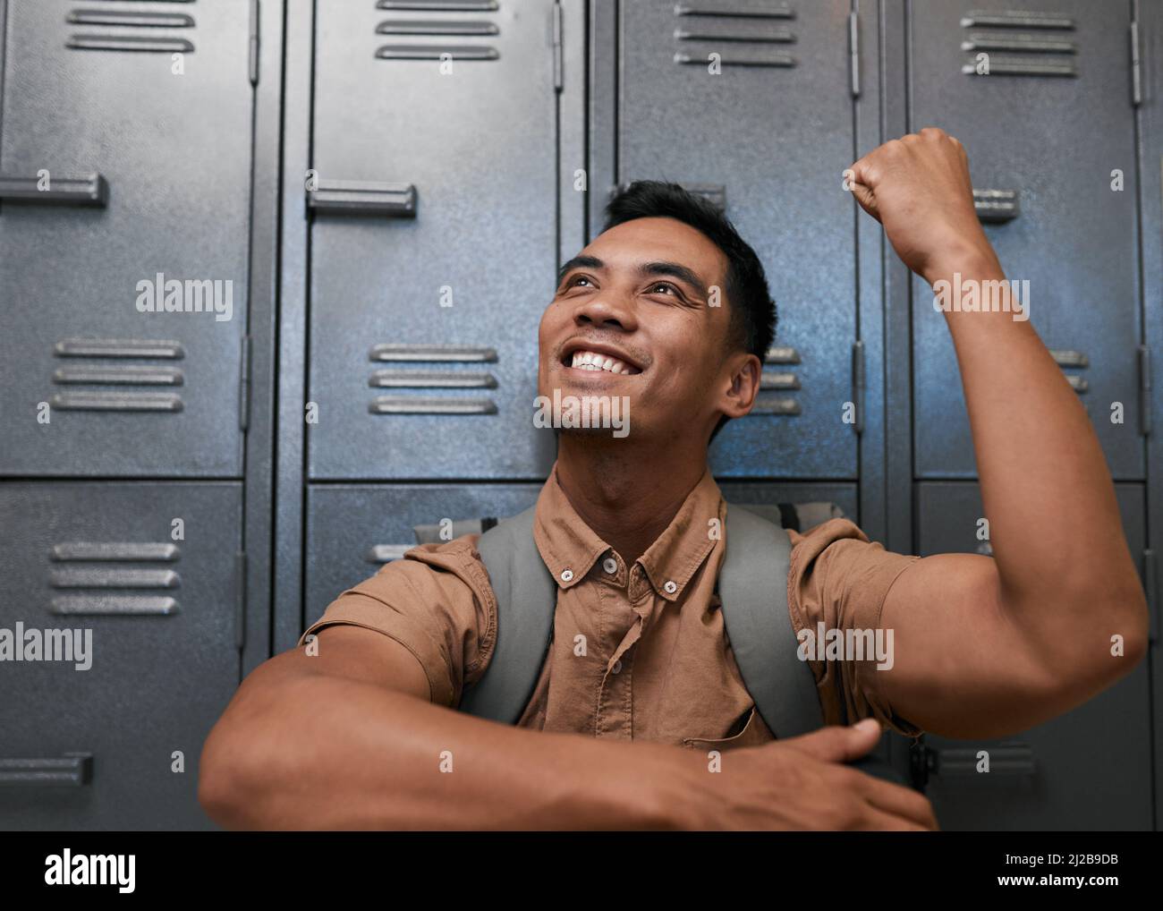 Ein junger südostasiatischer Student feiert vor den Schließfächern des Campus lächelnd Stockfoto