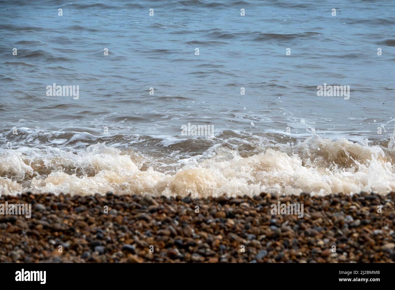 Kleine Wellen brechen an der Küste eines Kieselstrandes mit sprudelndem weißem Schaum und einem weichen Fokus Strand im Vordergrund Stockfoto