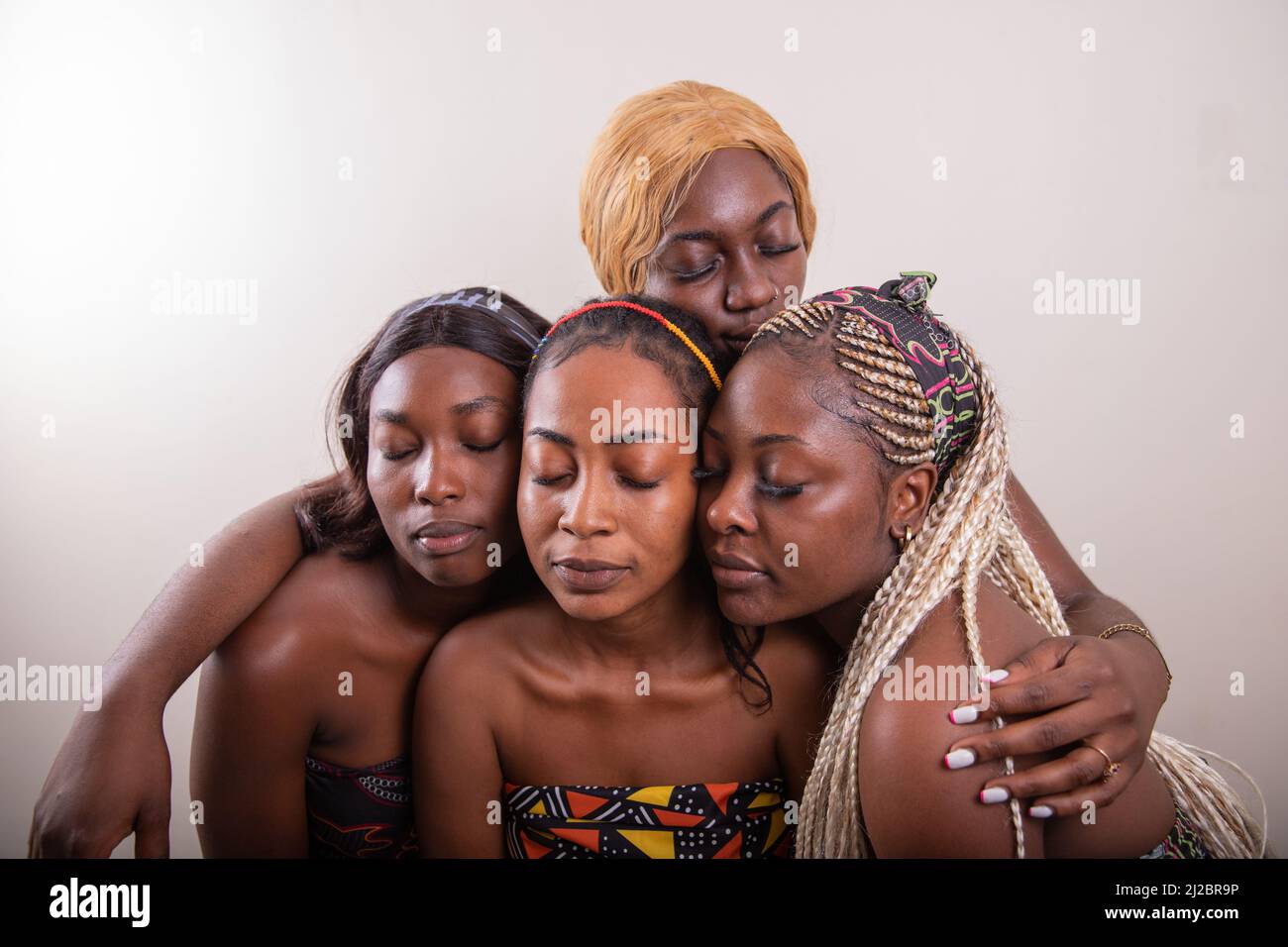 Vier afrikanische Mädchen umarmen sich im Studio in traditioneller Kleidung und schließen die Augen. Afrikanische Frauen vereint Stockfoto