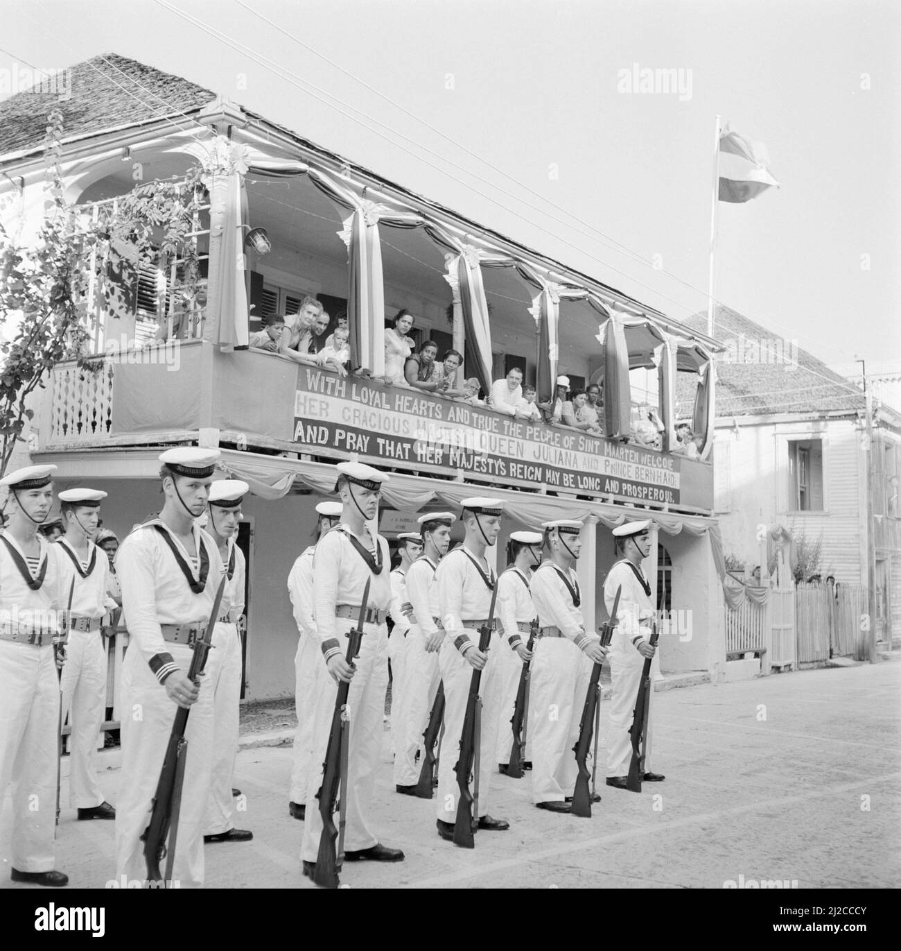 Seeleute warten in Philipsburg auf Sint Maarten vor einem Haus mit einem Banner. Die Inschrift lautet: Mit treuem Herzen und Treue empfangen die Menschen von Sint Maarten ihre gnädige Majestät Königin Juliana und Prinz Bernhard und beten, dass ihre Majestät lange und wohlhabend regieren möge um: 25. Oktober 1955 Stockfoto