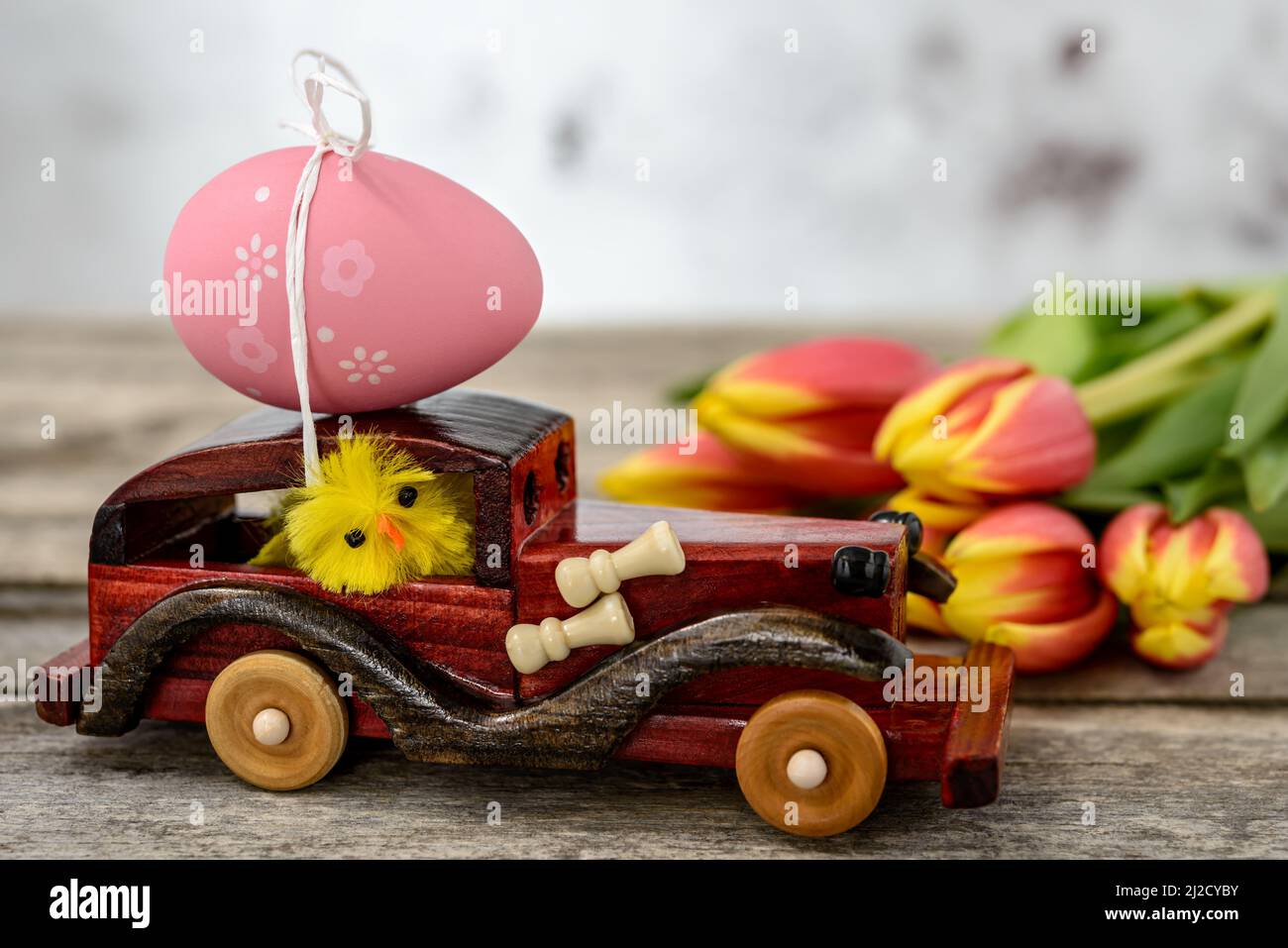 Ein Spielzeugauto aus Holz mit einem Osterei auf dem Dach und einem Hühnerfahrer neben einem Tulpenstrauß auf einem rustikalen Tisch. Stockfoto