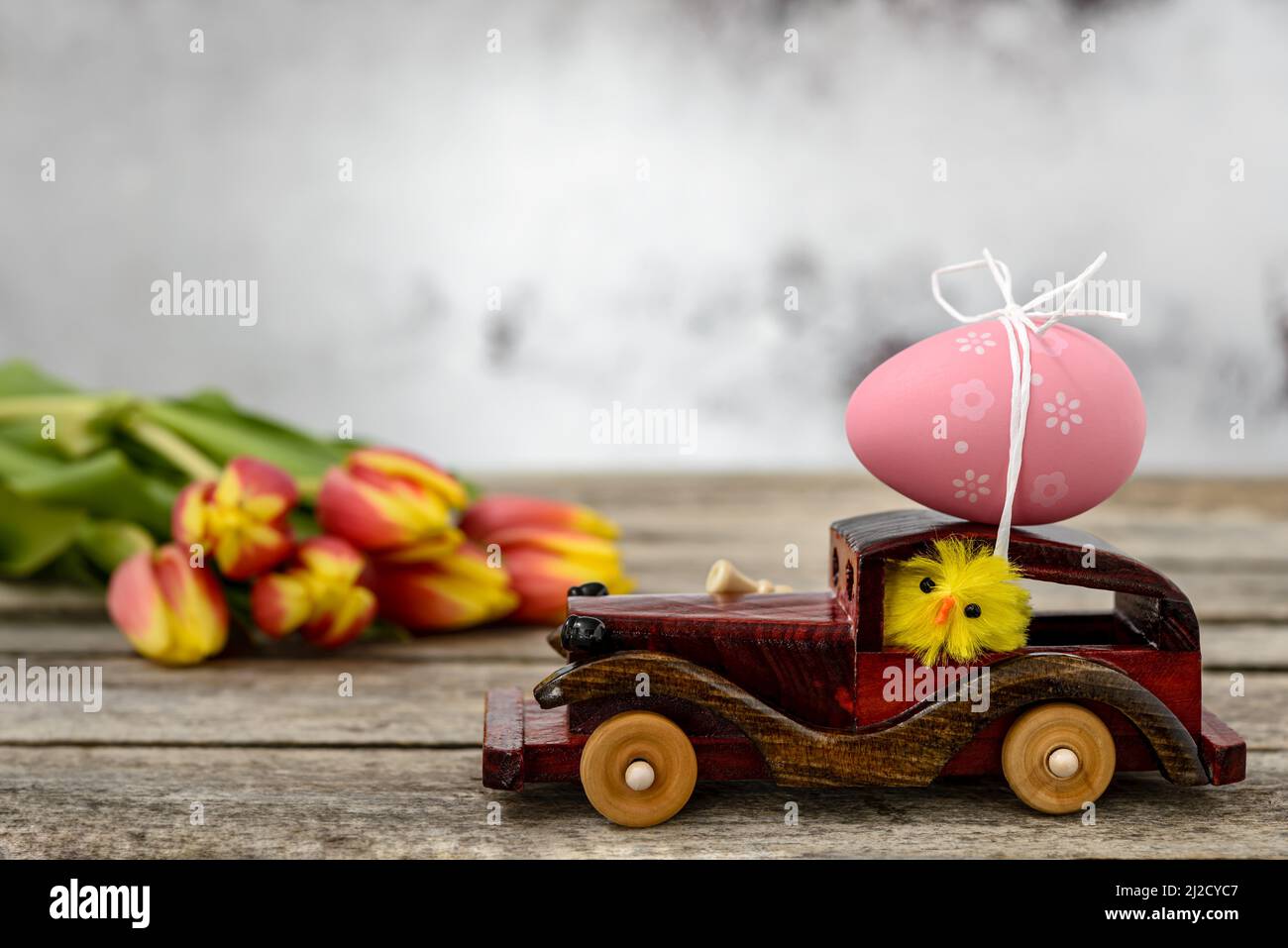 Ein Spielzeugauto aus Holz mit einem Osterei auf dem Dach und einem Hühnerfahrer neben einem Tulpenstrauß auf einem rustikalen Tisch. Stockfoto