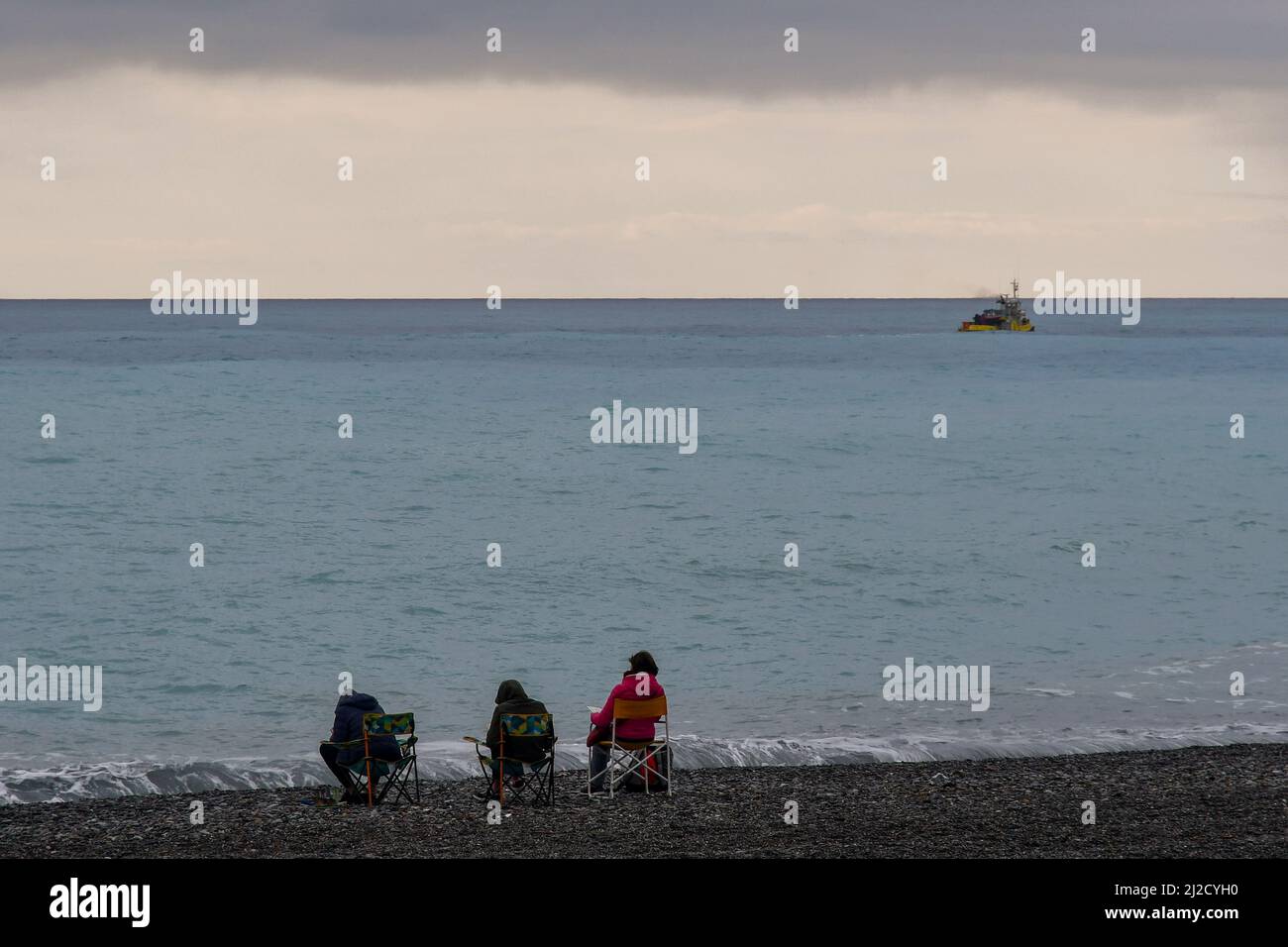 Drei Menschen von hinten sitzen am Strand am Meer mit einem Fischerboot an einem stürmischen Tag, Bordighera, Imperia, Ligurien, Italien Stockfoto