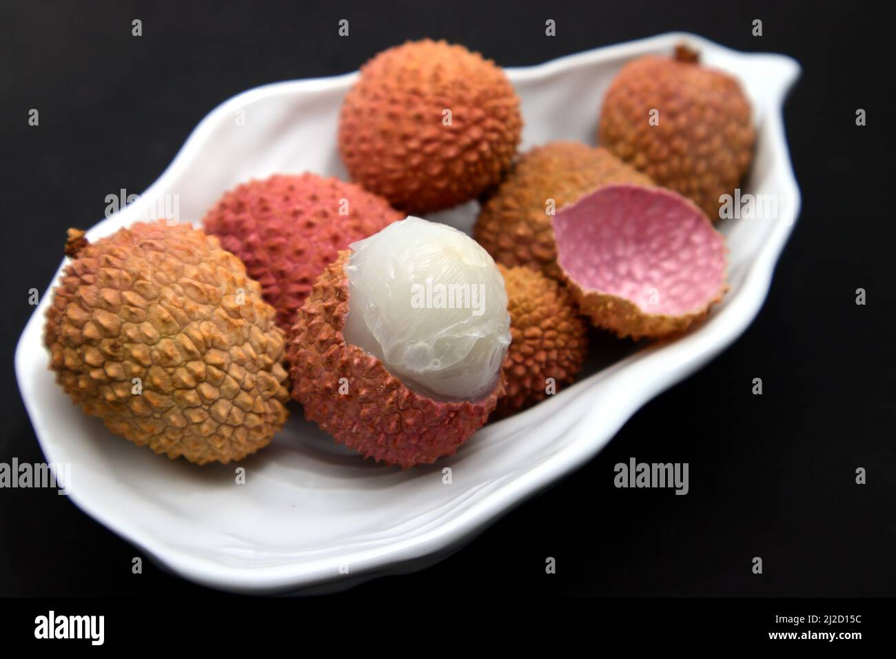 Nahaufnahme von geschälten und ungeschälten Litschis in einer weißen Platte auf einer schwarzen Oberfläche Stockfoto