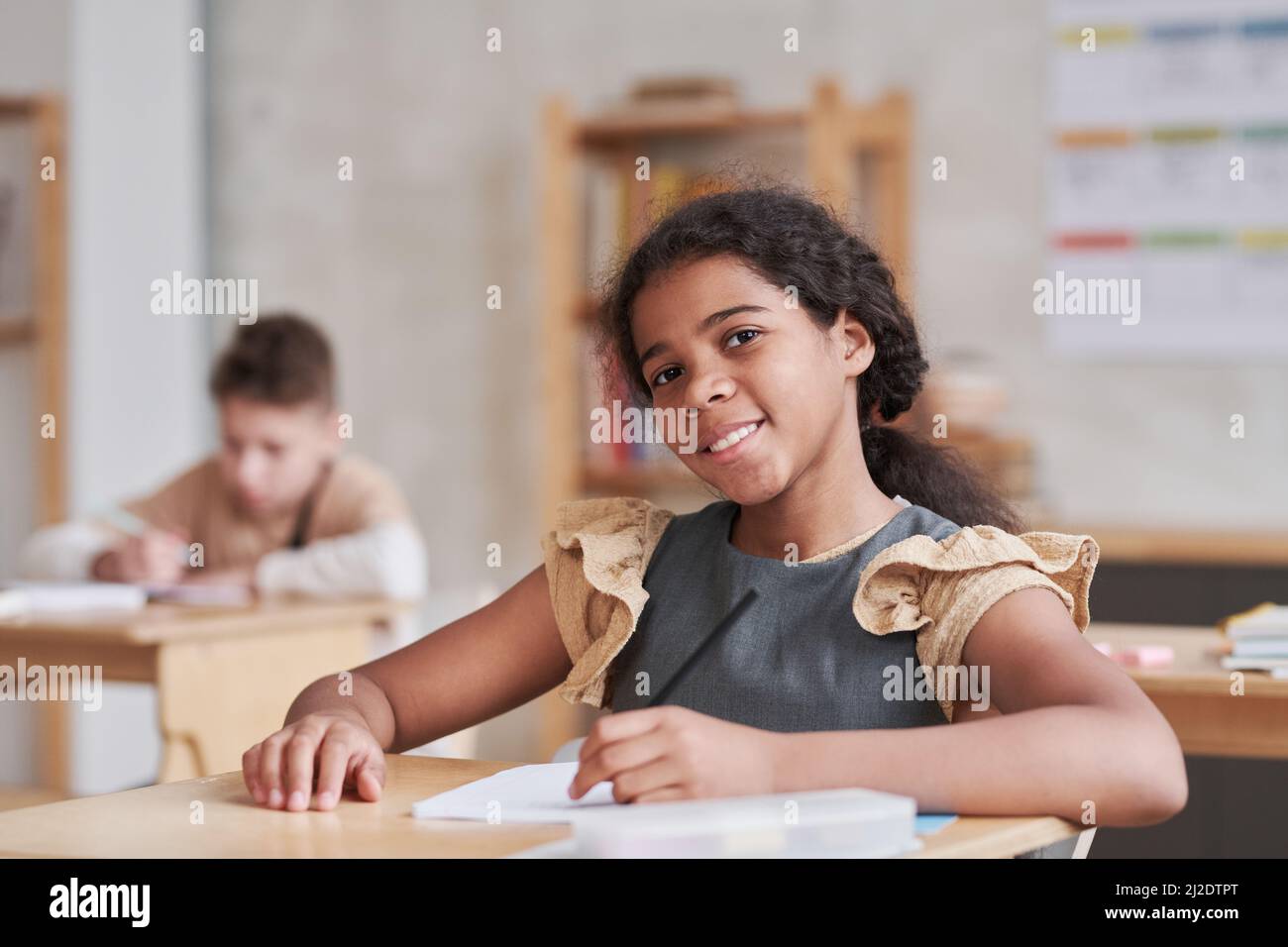 Porträt eines lächelnden Mädchens aus gemischter Rasse, das die Kamera anschaut, während es im Klassenzimmer am Schreibtisch sitzt, mit Holzdekor und Kopierraum Stockfoto