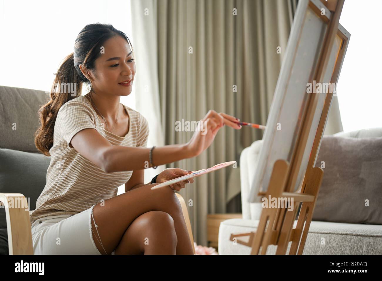 Eine inspirierte asiatische Frau malt im Wohnzimmer ein kreatives Kunstwerk auf eine LeinwandStaffelei mit Acrylfarbe. Stockfoto