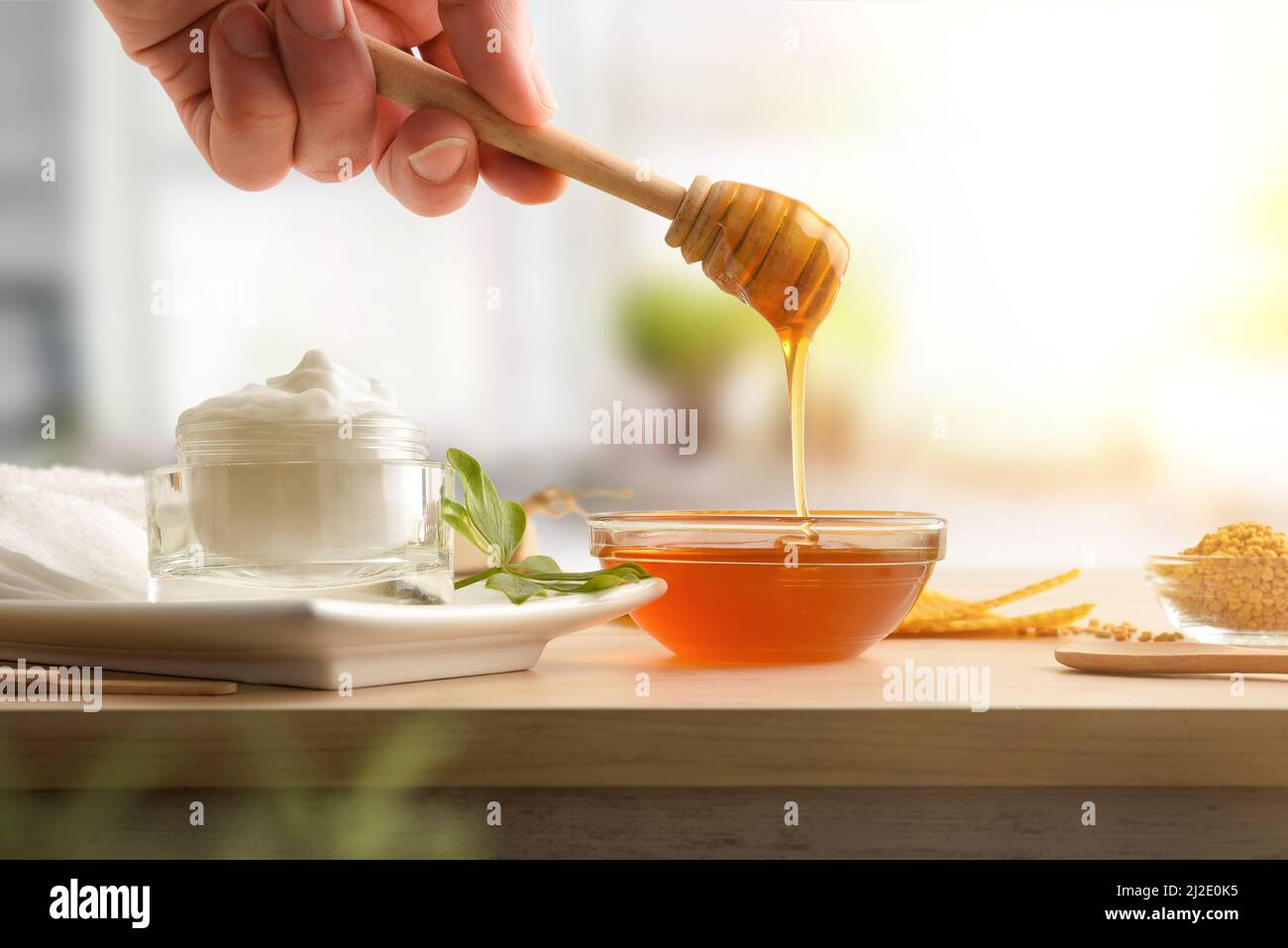 Glas mit Feuchtigkeitscreme für den Körper mit Honigextrakten und Hand mit Stick, der Honig, Pollen und Waben in einer Küche zeigt. Horizontale Zusammensetzung. Fr. Stockfoto