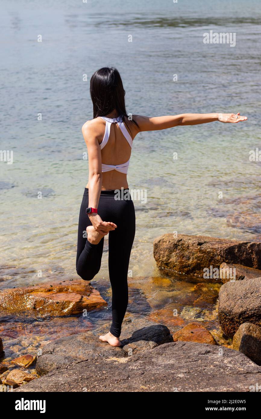 Eine schlanke Frau in schwarz-weißer Sportkleidung führt eine Quad-Stretch-Balance auf einem Bein auf Felsen am Strand durch. Im Hintergrund ist das Meer. Stockfoto