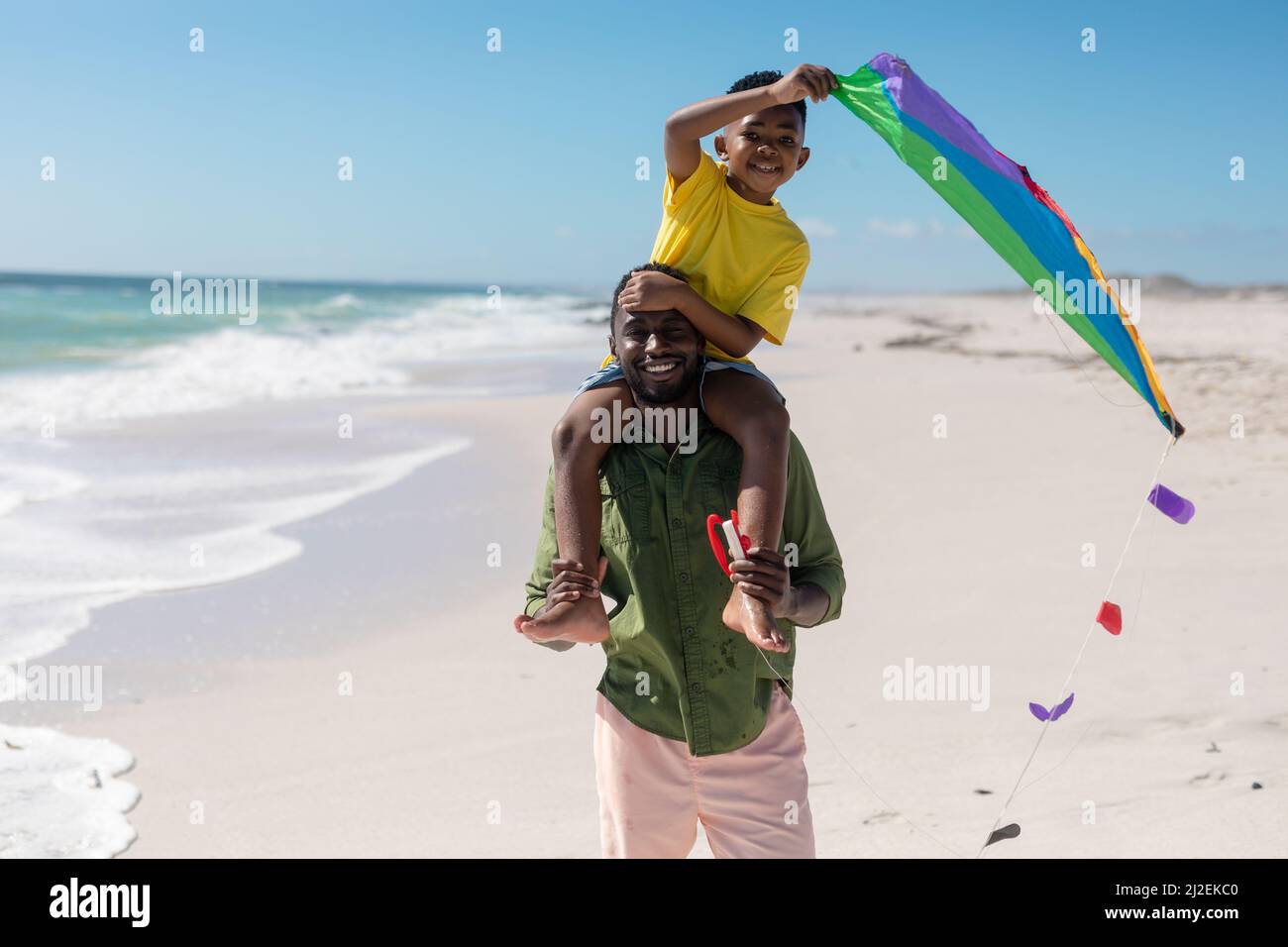 Porträt eines afroamerikanischen Mannes, der an sonnigen Tagen seinen Sohn mit Drachen auf den Schultern am Strand trägt Stockfoto