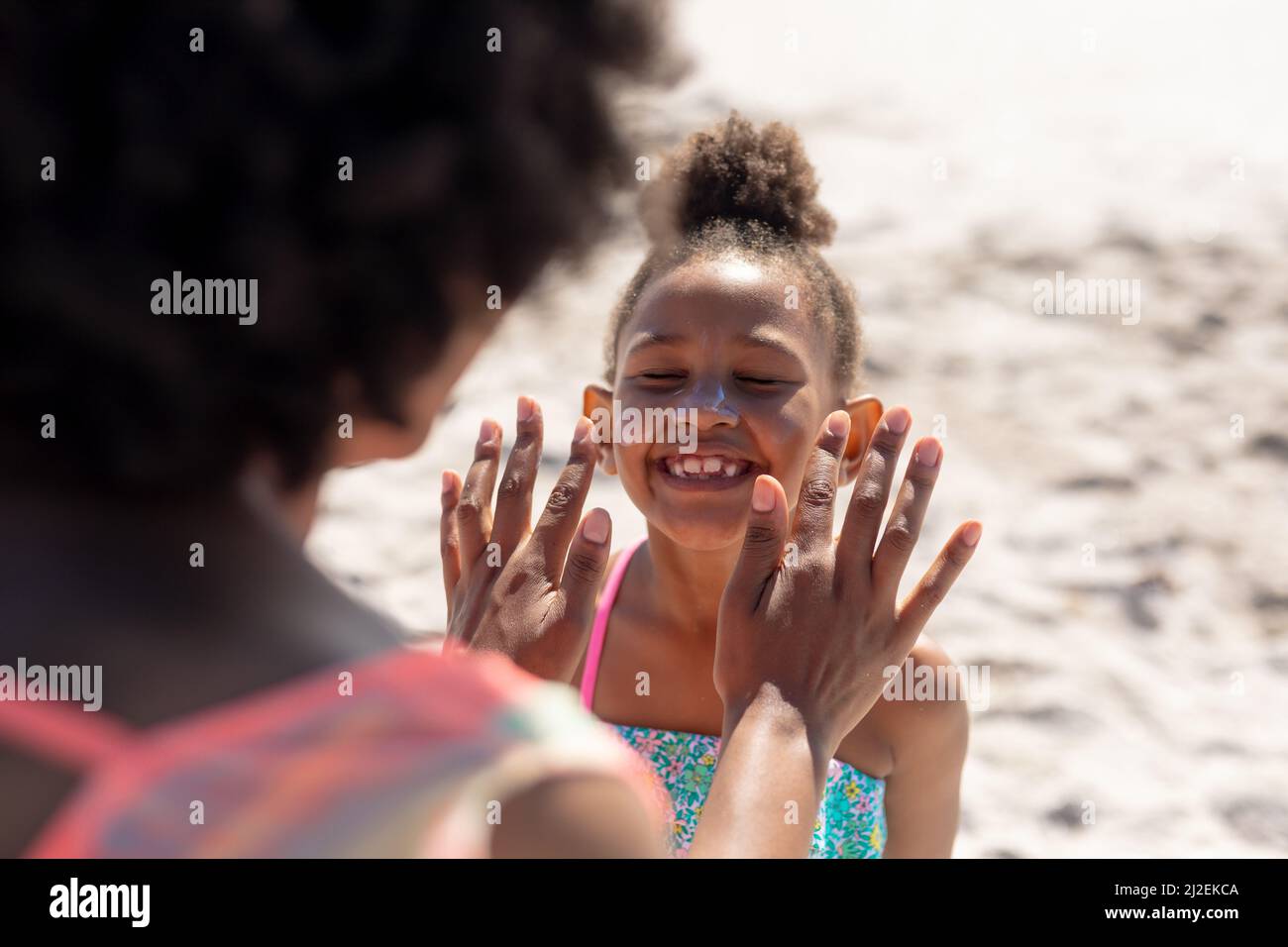 Fröhliches afroamerikanisches Mädchen lächelt mit geschlossenen Augen, während die Mutter Sonnencreme auf das Gesicht aufgibt Stockfoto