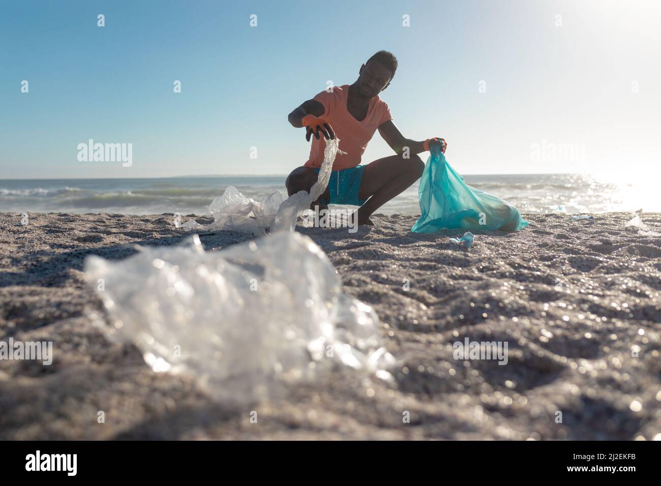 afroamerikanischer Mann hocke beim Sammeln von Müll in Plastiktüte am Strand an sonnigen Tag Stockfoto