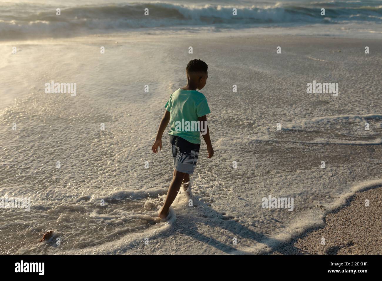 Lange Rückansicht des afroamerikanischen Jungen, der an sonnigen Tagen am Strand an der Küste watend war Stockfoto