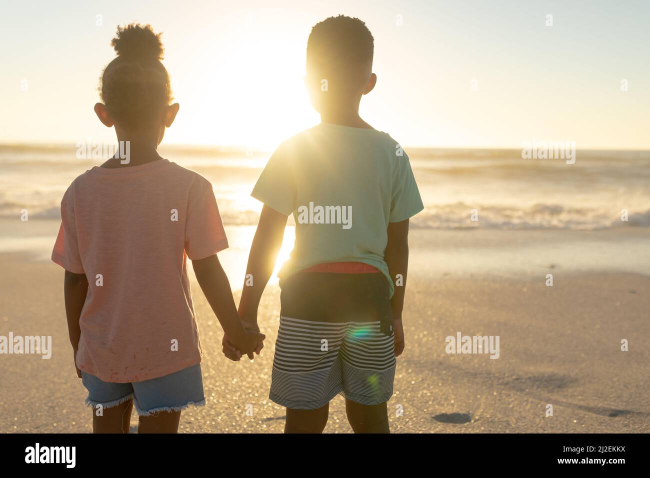 Rückansicht von afroamerikanischen Geschwistern, die sich die Hände halten, während sie vom Strand aus den Sonnenuntergang über dem Meer beobachten Stockfoto