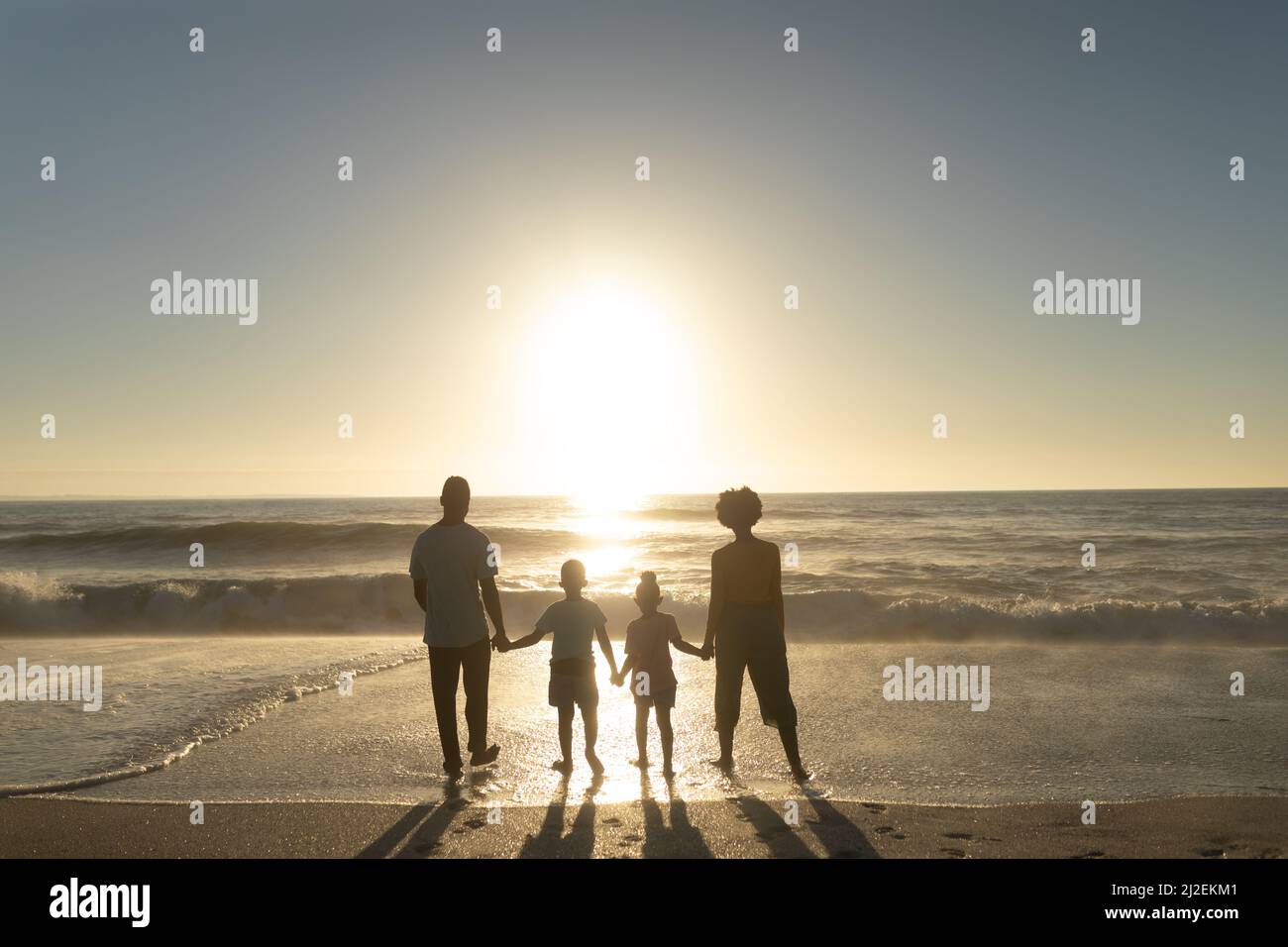 Lange Rückansicht der afroamerikanischen Familie, die sich die Hände hält, während sie den Sonnenuntergang über dem Meer betrachtet Stockfoto