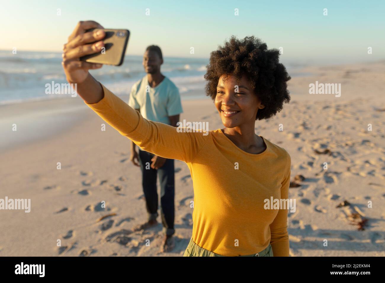 Glückliche afroamerikanische Frau, die Selfie mit einem Mann macht, der im Hintergrund am Strand steht Stockfoto