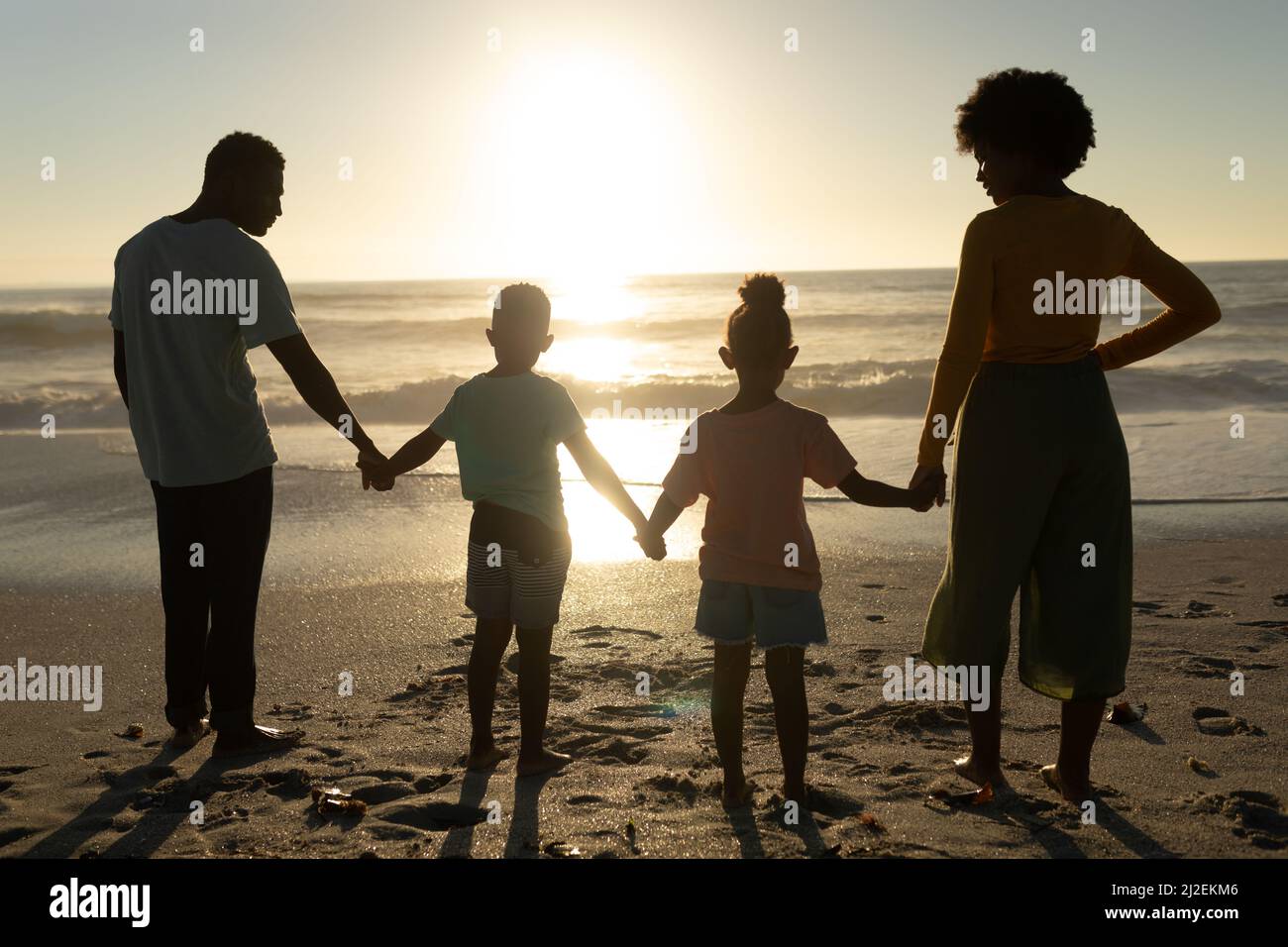 Rückansicht einer afroamerikanischen Familie, die sich die Hände hielt, während sie vom Strand aus den Sonnenuntergang über dem Meer betrat Stockfoto