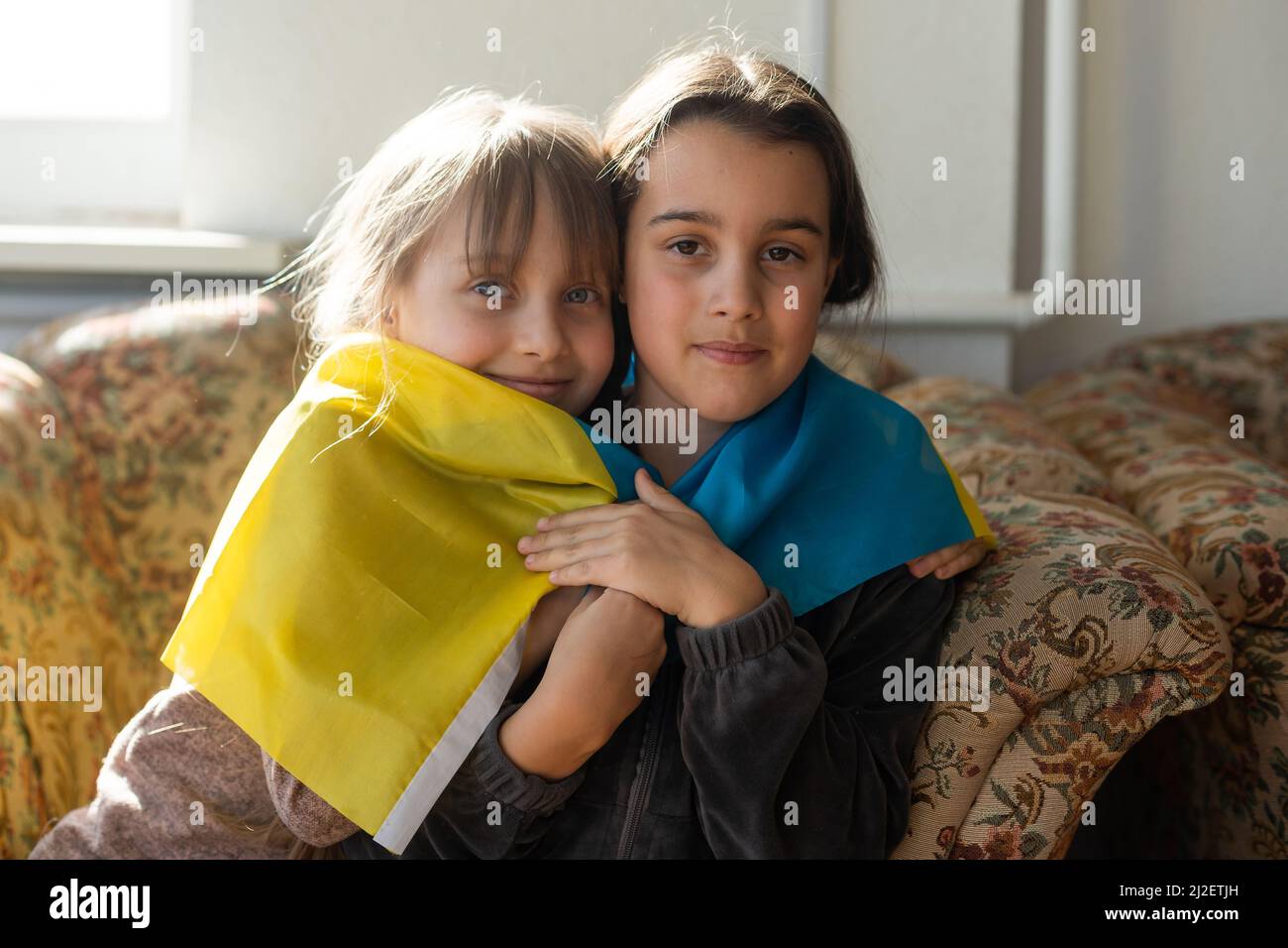 Kleine Mädchen mit ukrainischer Flagge vor einem Fenster. Die kleinen Mädchen winken die Nationalflagge, während sie für den Frieden beten Stockfoto