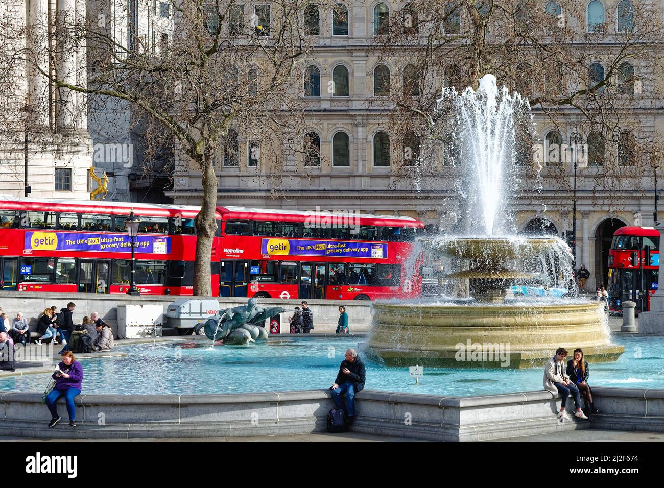 Menschenmengen, die an einem der Brunnen am Trafalgar Square mit roten Londoner Bussen im Hintergrund an einem sonnigen Frühlingstag im Zentrum von London, England, sitzen Stockfoto