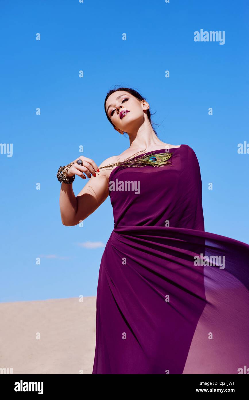 Erstaunlich schöne Brünette Frau mit der Pfauenfeder in lila Stoff in der Wüste. Orientalisches, indisches, Mode- und Stilkonzept Stockfoto