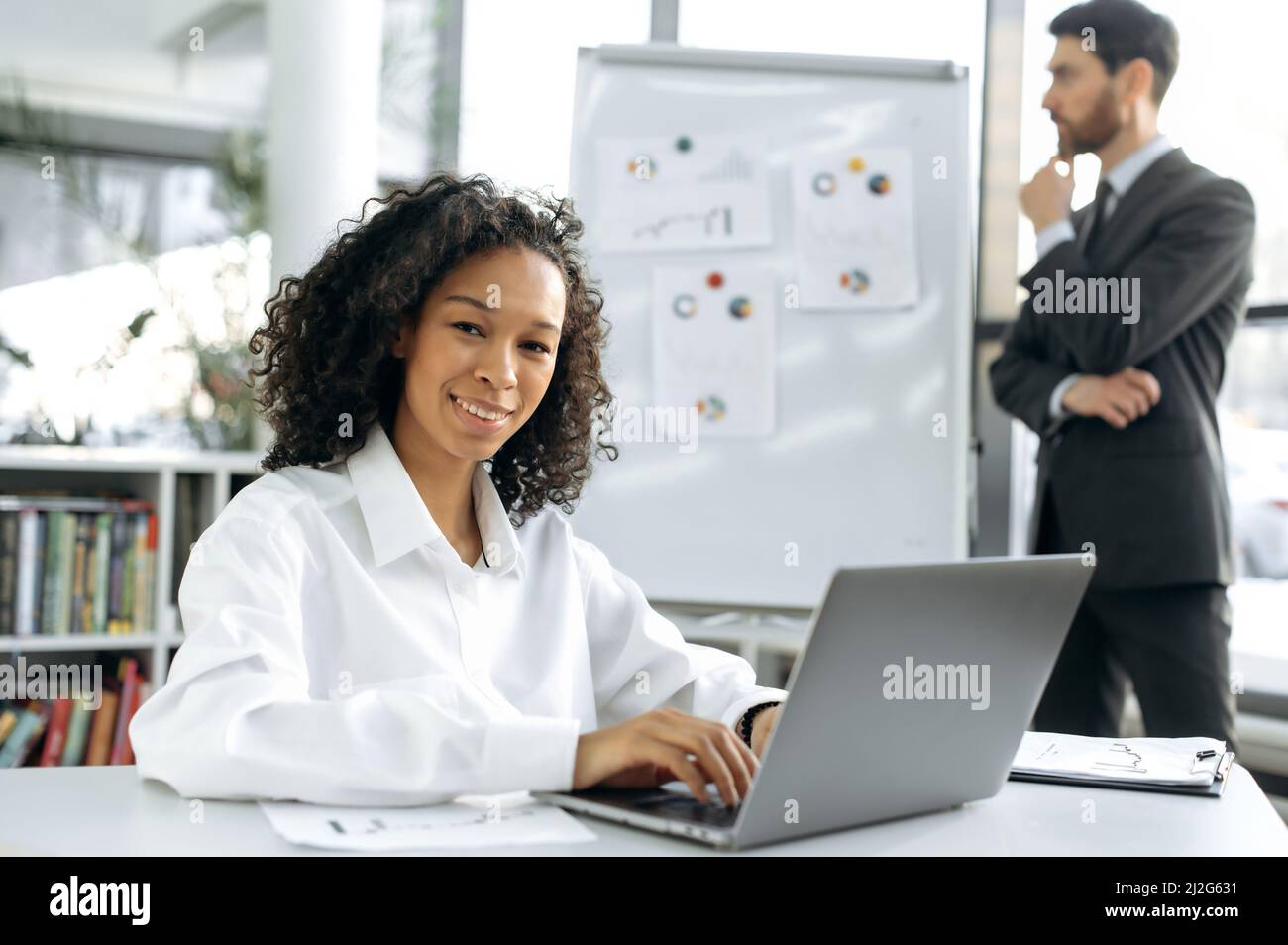 Die erfolgreiche afroamerikanische Frau sitzt an ihrem Schreibtisch vor einem Laptop, schaut lächelnd auf die Kamera. Im Hintergrund arbeitet ein männlicher Kollege mit Diagrammen, entwickelt eine Strategie zur Modernisierung der Arbeit Stockfoto
