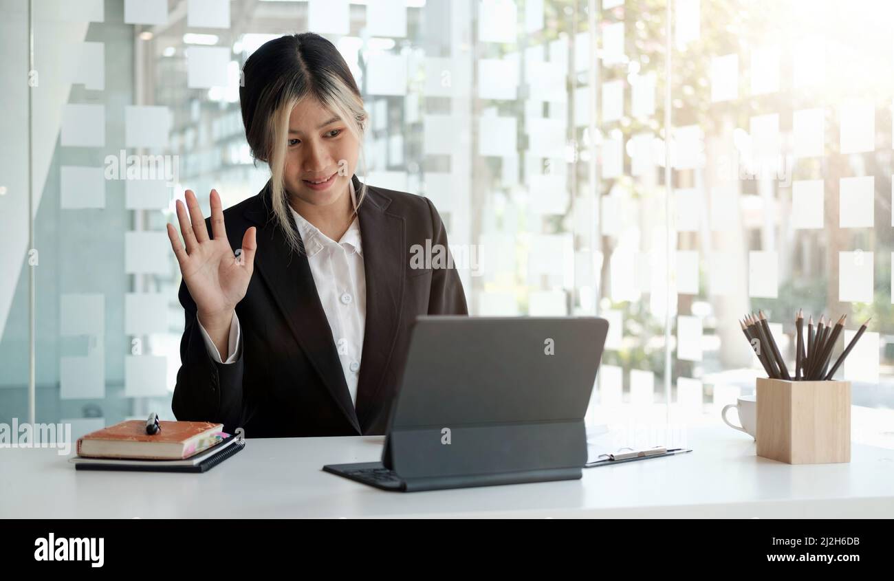 Junge asiatische Frau im Anzug lächelnd und winkende Hand auf Laptop, während sie im Büro sprach oder chattet. Stockfoto
