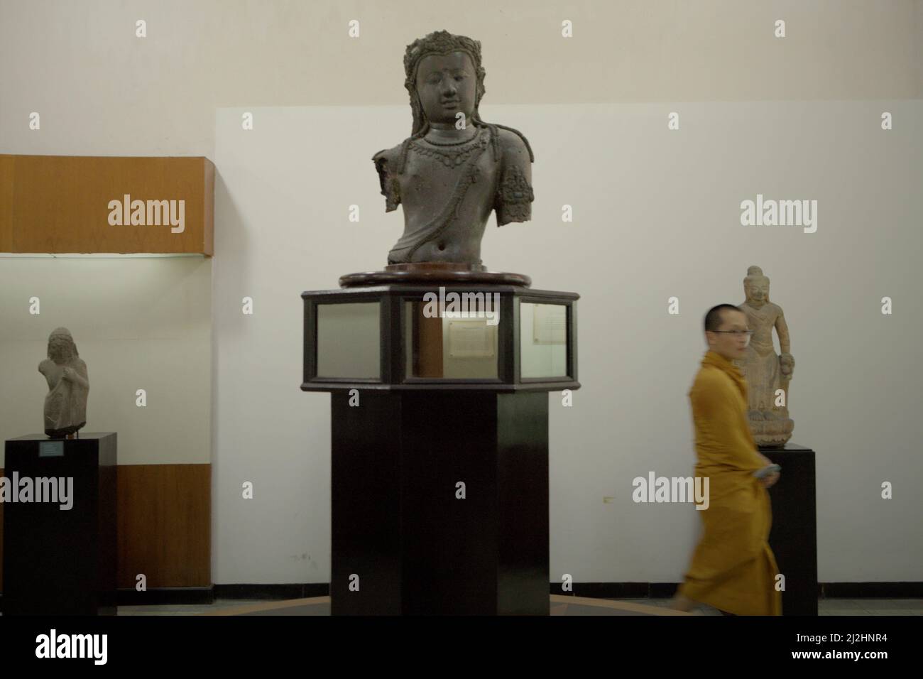 Ein Mönch, der hinter einer Srivijaya-Skulptur von Bodhisattva Avalokiteswara aus der Srivijaya-Zeit im Nationalmuseum, Bangkok, Thailand, vorbeikommt. Die ikonische Skulptur, die in der Provinz Surat Thani im Süden Thailands gefunden wurde, ist in Form von Repliken in Chaiya, einer Stadt in derselben Provinz, so vertraut. Es wird angenommen, dass 'Chaiya' von 'Srivijaya' oder 'Sri Vichai' oder 'Siwichai' im lokalen Dialekt abgeleitet wird. Stockfoto