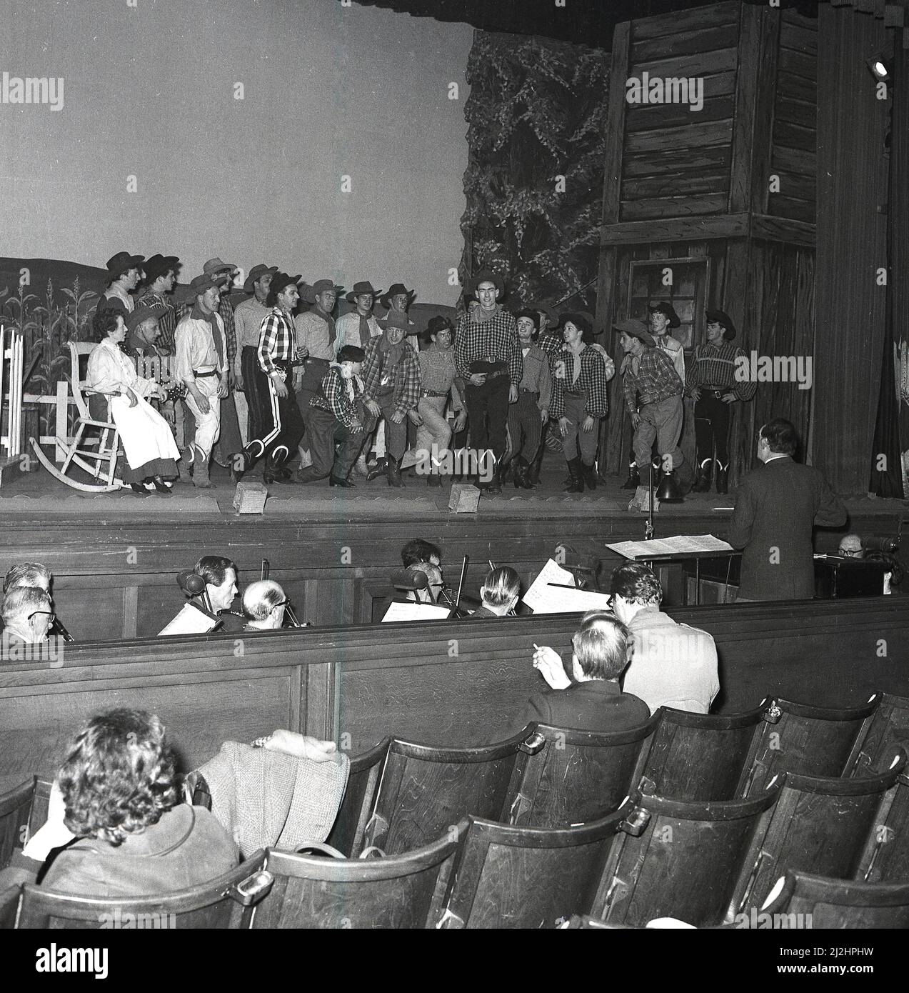 1960s, historische, große Gruppe von Schauspielern auf einer Bühne während einer Aufführung des amerikanischen Musicals Oklahoma, produziert von der Kelty Musical Association in der Carnegie Hall, Dufermline, Fife, Schottland, Großbritannien, Mit Orchester in der Grube. Oklahoma basiert auf dem Theaterstück „Green Grow The Lilacs“ aus dem Jahr 1931 und ist das erste Musical von Rodgers und Hammerstein, das 1943 am Broadway eröffnet wurde. Basierend auf den Romanzen eines einfachen Bauernmädchen ist das Musical eine beliebte Wahl für Schulen und Gemeinschaftsgruppen. Stockfoto