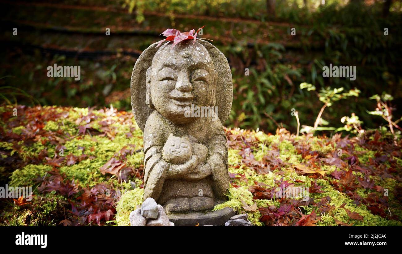 Nahaufnahme einer Steinstatue eines lächelnden Jizo Bosatsu, der eine Katze im Park hält Stockfoto