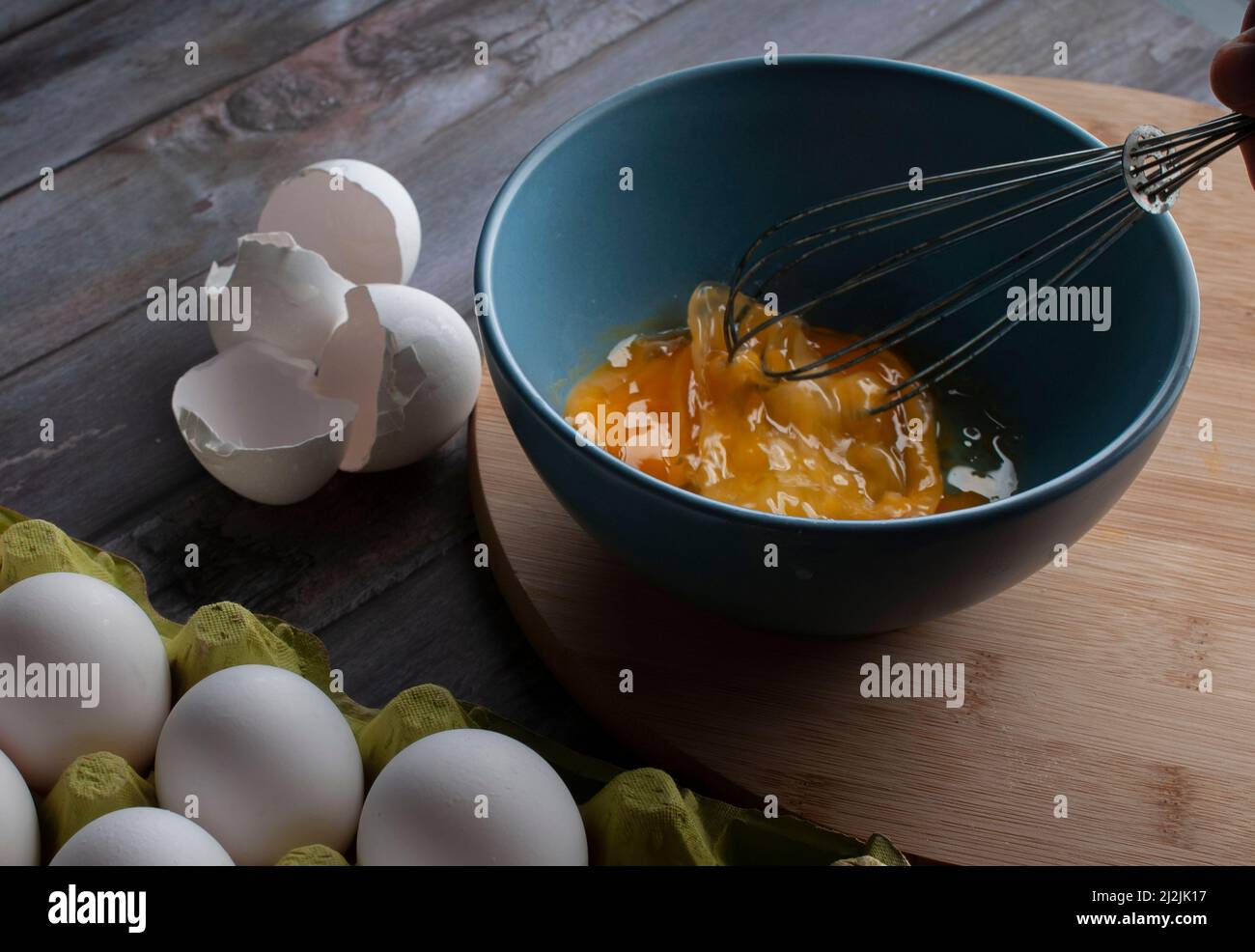 Rührei roher Eier in einer blauen Schüssel über einem runden Holzbrett und Hände, die die Eier mischen, Stockfoto