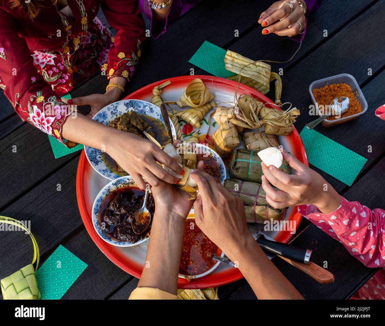 Draufsicht auf die Hände der muslimischen Familie, die am Ende des Ramadan Mubarak auf einem runden Tablett auf einem schwarzen Tisch eine Vielzahl traditioneller malaiischer Gerichte genießen Stockfoto
