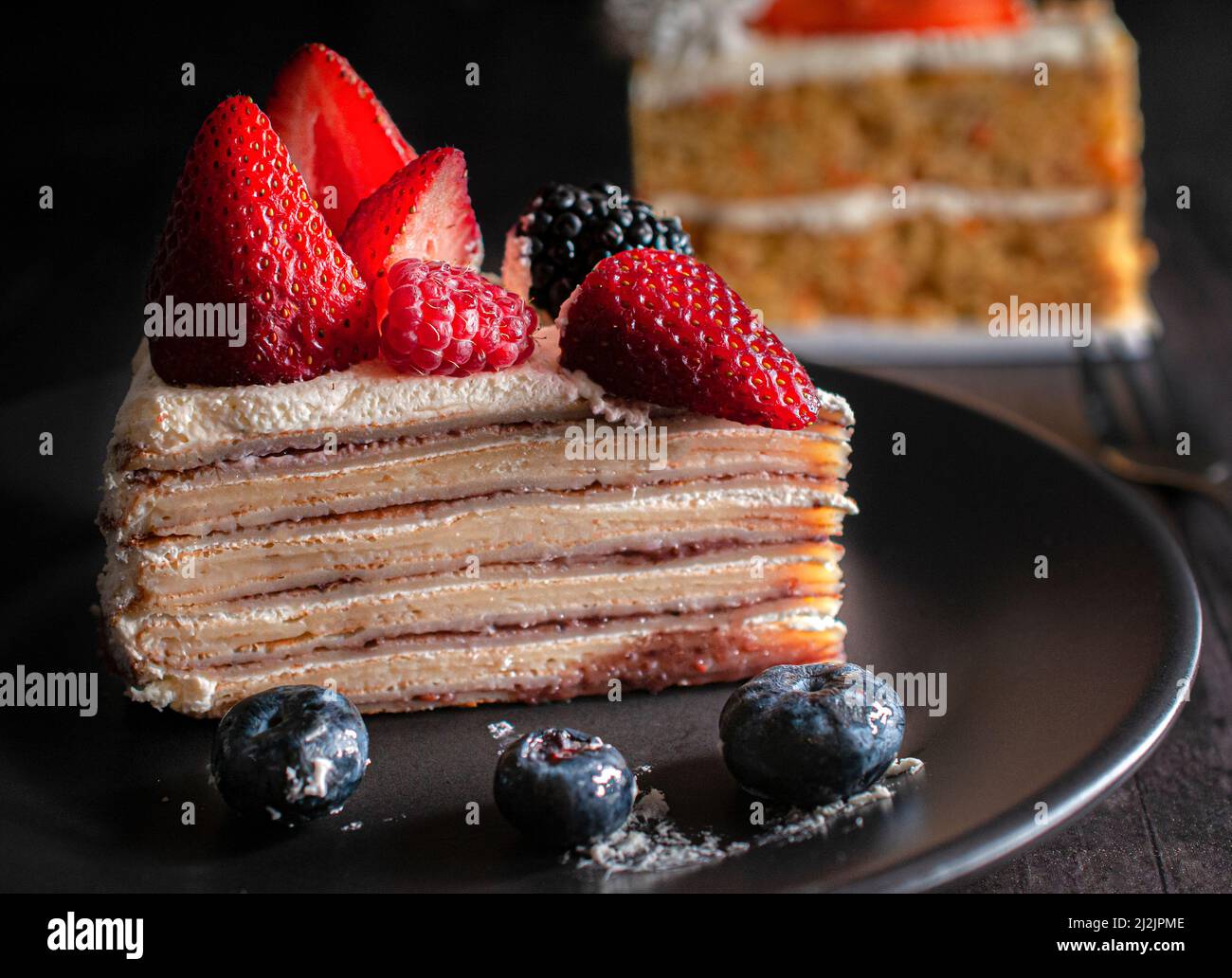 Scheibe Bananenkrepskuchen mit Creme und Erdbeeren oben mit blauen Beeren auf einem schwarzen Teller. Makro, dunkle Food-Fotografie. Stockfoto