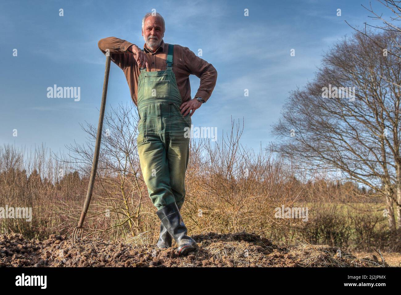 Ein Bauer in grünen Latzhose und Gummistiefeln steht auf einem Misthaufen und hat sich an seine Mistgabel gelehnt. Stockfoto