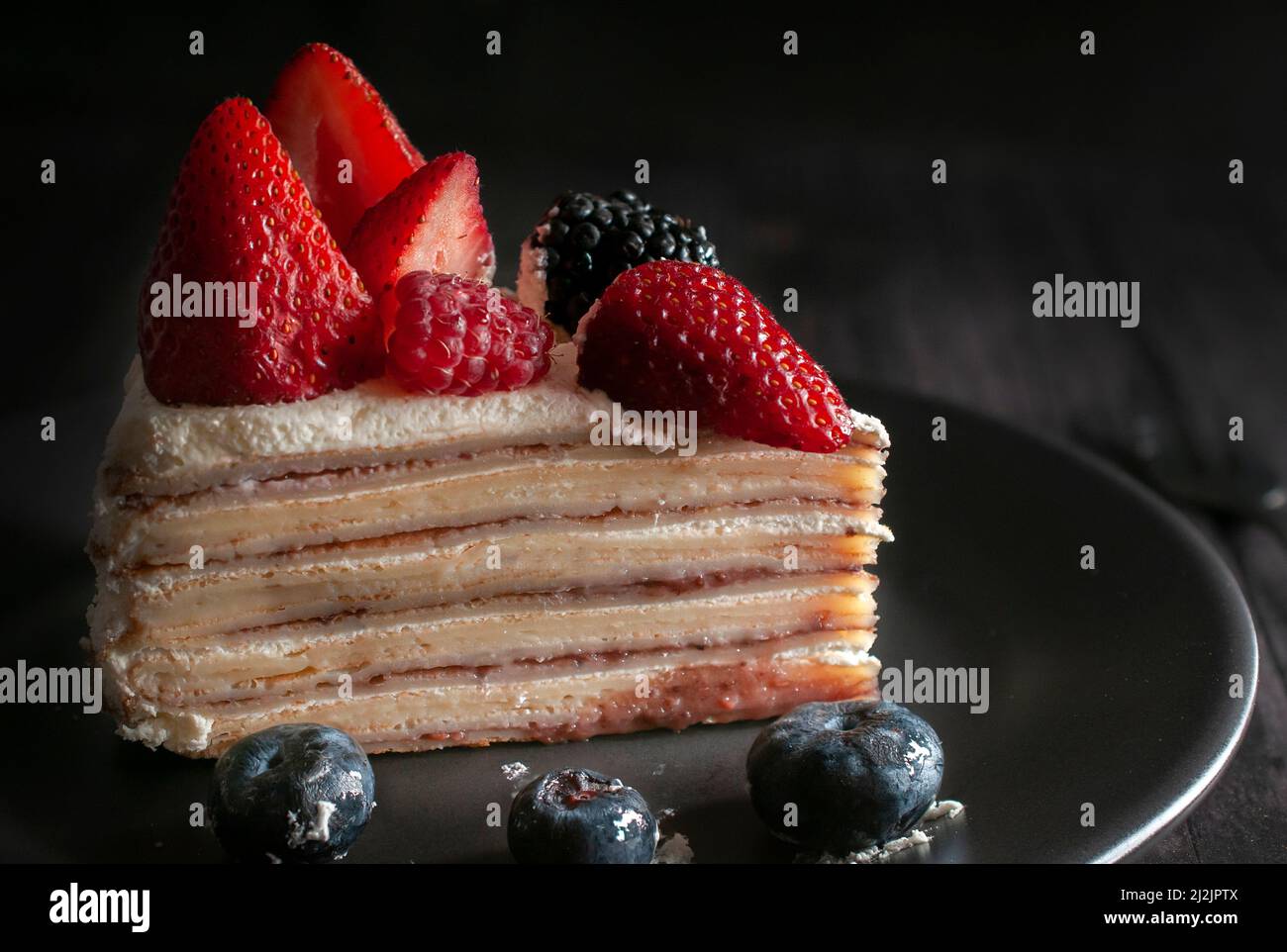 Scheibe Bananenkrepskuchen mit Creme und Erdbeeren oben mit blauen Beeren auf einem schwarzen Teller. Makro, dunkle Food-Fotografie. Stockfoto