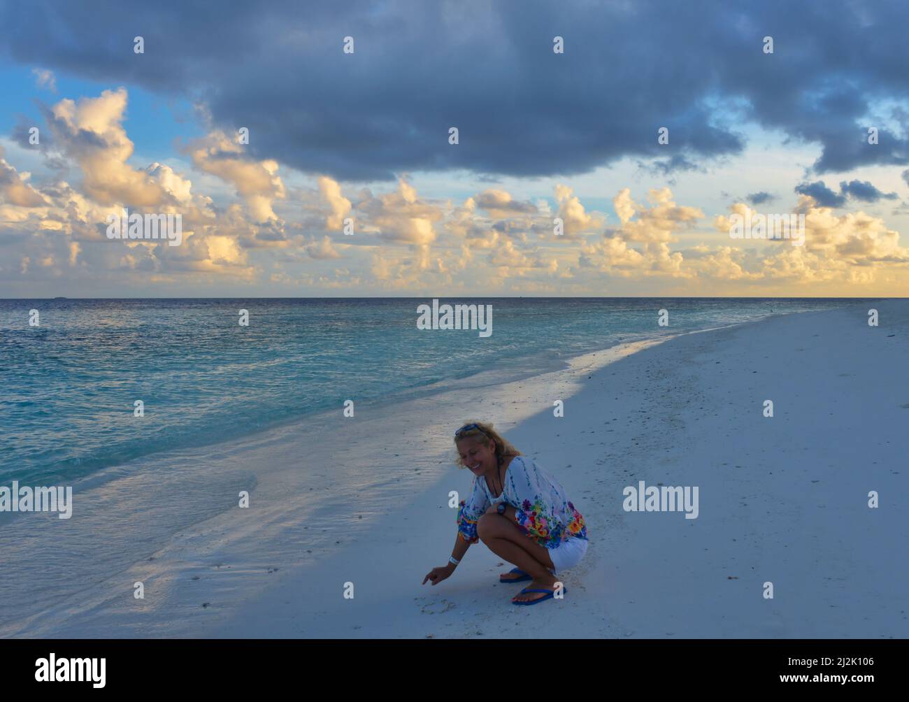 Lächelnde Frau, die beim Sonnenaufgang am Strand hockend im Sand zieht, Malediven Stockfoto