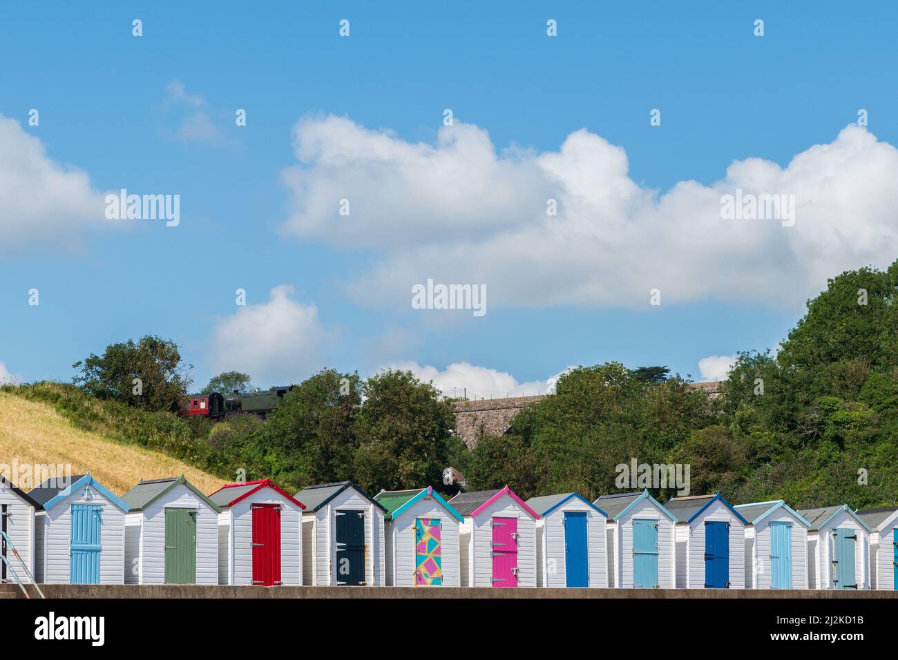 Farbenfrohe Strandhäuser. Eine Reihe von bunten Strandhütten mit einer dampfenden Dampfeisenbahn auf einem Steinviadukt gegen den blauen Himmel. Stockfoto