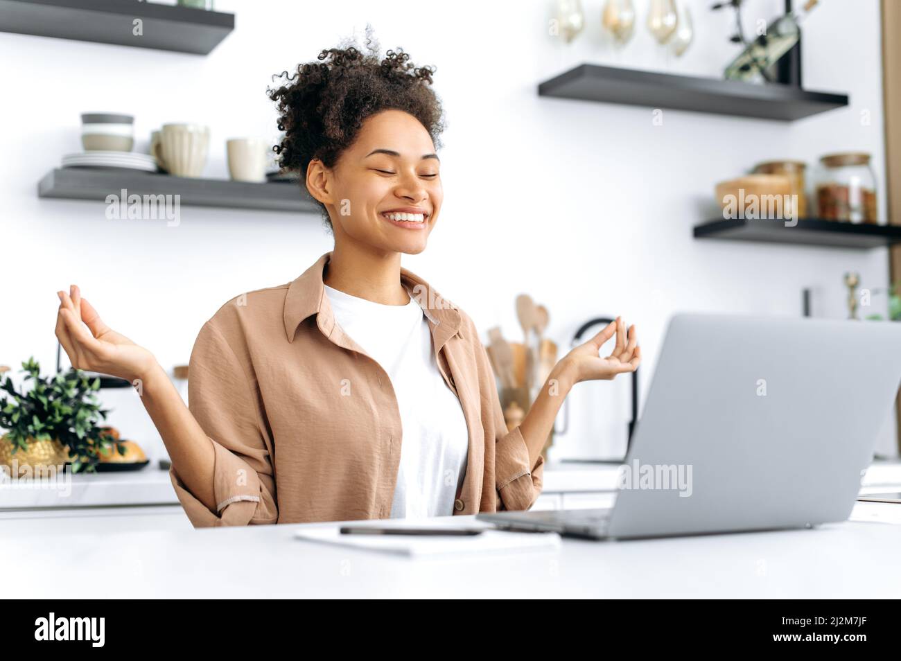 Hübsche, positive, ruhige, entspannte afroamerikanische junge Frau, Studentin, Freiberuflerin oder Designerin, die fernab von zu Hause an einem Laptop arbeitet, sich ausruhen und meditieren während der Arbeit, geschlossene Augen, ruhiges Konzept Stockfoto