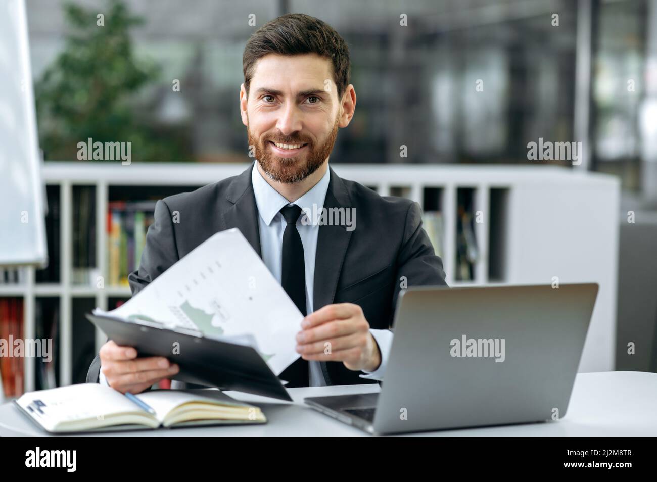 Der attraktive kaukasische Manager für gewerbliche Immobilien, Manager, sitzt an einem Schreibtisch vor einem Laptop, in einem modernen Büro, in stilvoller Business-Kleidung, arbeitet mit Dokumenten, blickt auf die Kamera und lächelt Stockfoto