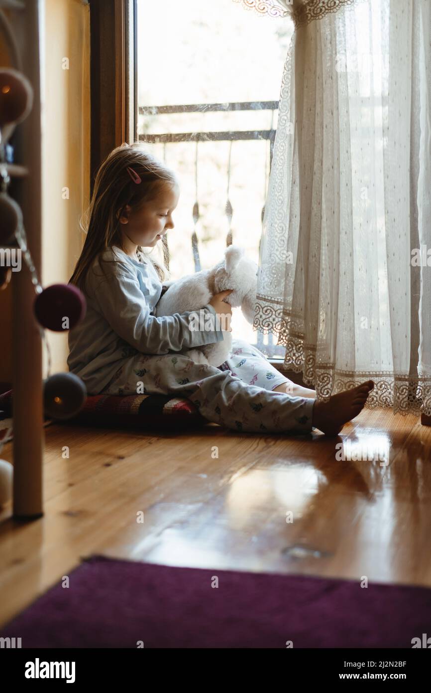 Kind sitzt auf dem Boden und spielt Ameise nach Hause. Verträumte, sonnige Stimmung. Mädchen, das von hinten durch das Fenster schaut. Niedliches Kind wartet darauf, spazieren zu gehen Stockfoto