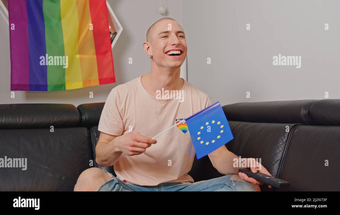 Der kaukasische schwule Mann winkt mit der europäischen Flagge, sitzt auf einem Sofa, lächelt und hält die Fernbedienung. Hochwertige Fotos Stockfoto