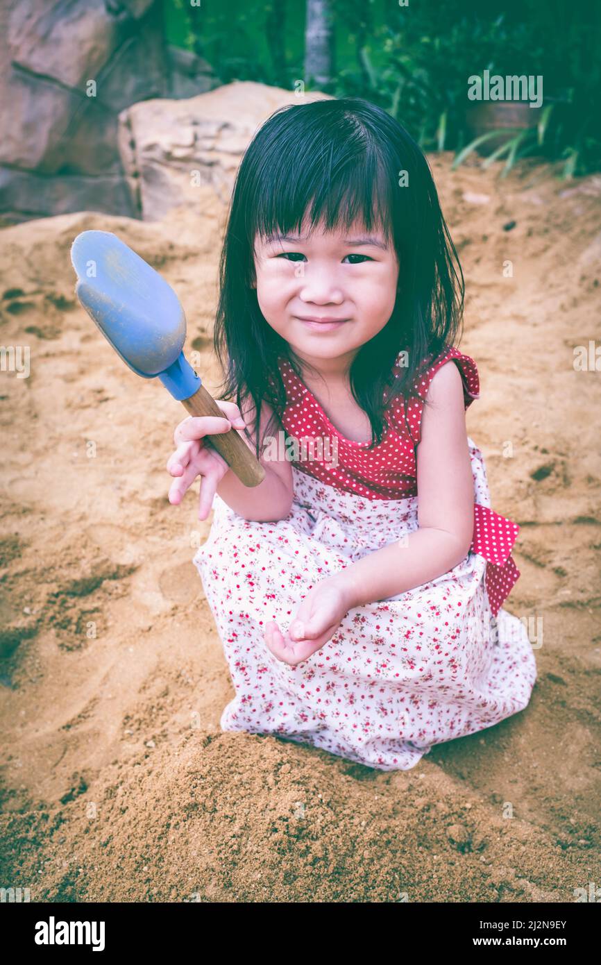Glückliches Kind, das mit Sand spielt und lächelt. Liebenswert asiatische Mädchen hat Spaß Graben in den Sand an einem Sommertag und Blick auf die Kamera. Vintage-Ton. Outdoo Stockfoto