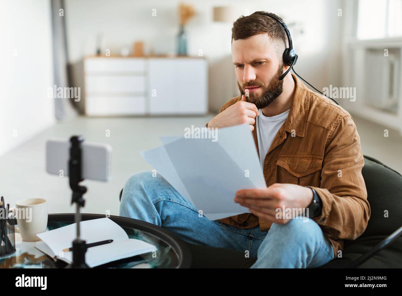 Nachdenkliche Männliche Lesepapiere Making Video Auf Smartphone Zu Hause Stockfoto