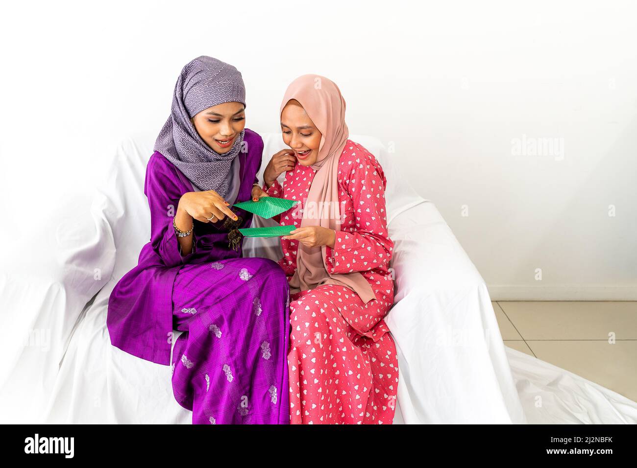 Zwei muslimische Mädchen, die in purpurroten und roten Kebaya-Trachten gekleidet sind, tauschen Geldpaketgeschenke aus, um das Ende des Ramadan zu feiern und sitzen auf dem weißen Sofa Stockfoto